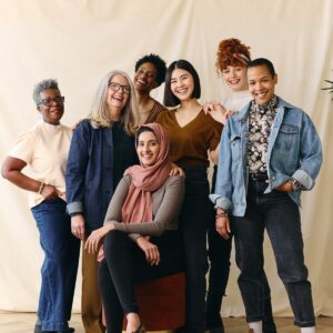 A group of women stand and sit around a chair with a beige background behind them. 