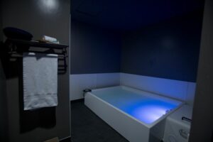 Image of rectangular bath tub in a dark room with dark wallpaper. There is a blue light in the water and on a wall on the left hand side there is a black towel rack with a white towel hanging from it. The image is of a float spa. 