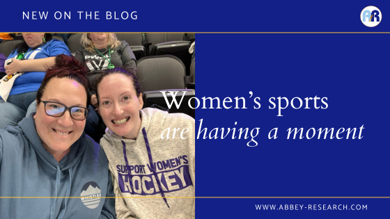 Blog header with the title "women's sports are having a moment." The picture is of two White women, smiling at the camera. The one on the left has red hair with black glasses and a blue hoodie, and the one on the right has a cream colored hoodie that says "Support Women's Hockey" and has dark brown hair. 
